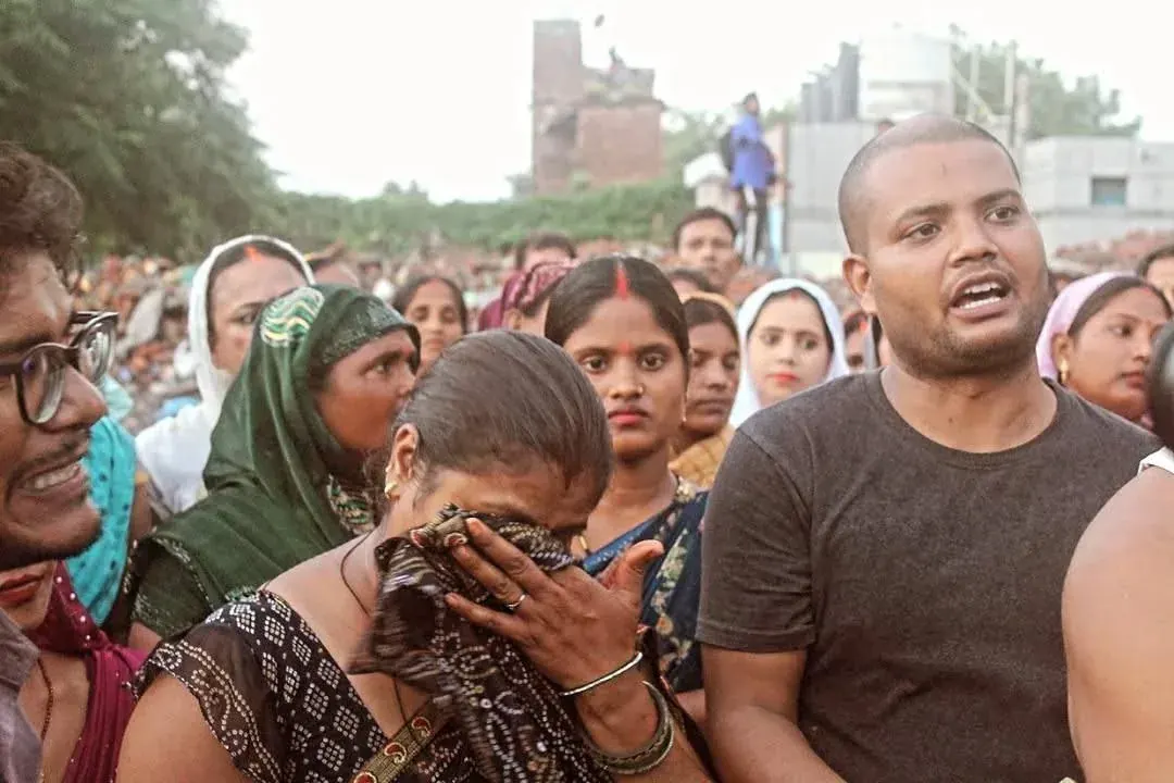 Group of people outdoors, including a woman covering her face with a scarf and a man speaking, suggesting a public discussion or gathering