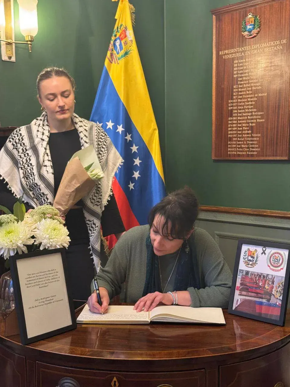 Two women indoors near a flag and a plaque, one seated signing a book and the other holding flowers